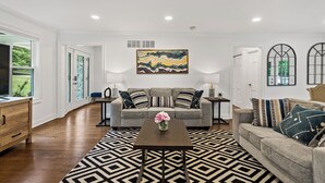 A living room with a sofa, coffee table, and decorative pillows. There is a painting on the wall and a window with a view of greenery. - Estate 5BR 3.5Bath with IGLOO, Game Rm, Fire Pit sleeps 16- 5min Princeton dwtn (Princeton)