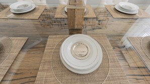 Mesa de jantar com tampo de vidro, rodeada por cadeiras brancas, com pratos e jogos americanos.