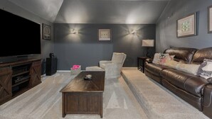 Sala de estar com televisão de tela plana, sofá de couro marrom, poltrona branca, mesa de centro de madeira e duas luminárias fixadas na parede.