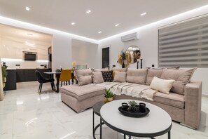 A modern living room with a sofa, ottoman, and coffee table, a dining area with chairs and a table, and a kitchen with a TV and counter.