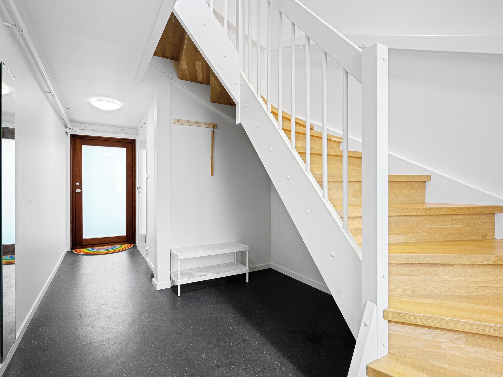 Un escalier moderne avec des marches en bois et des rampes blanches, un banc et une porte dotée d’un panneau de verre.