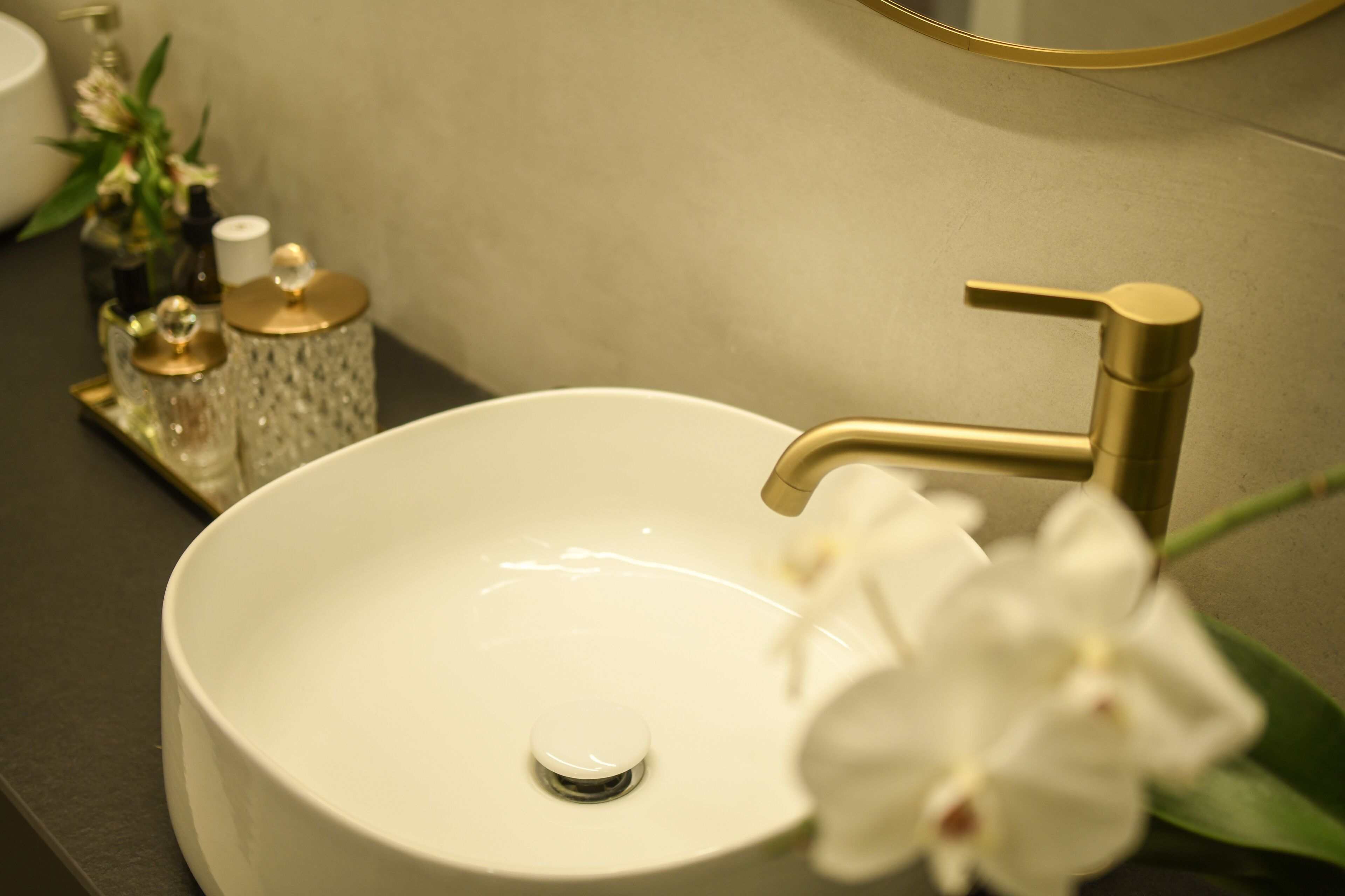 A white sink with a gold faucet, a small mirror above it, and a vase with flowers on the counter.