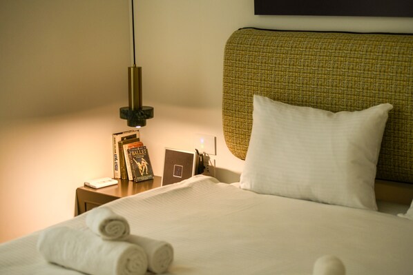 Een netjes opgemaakt bed met opgerolde handdoeken, een nachtkastje met boeken en een lamp, en een wandlamp.