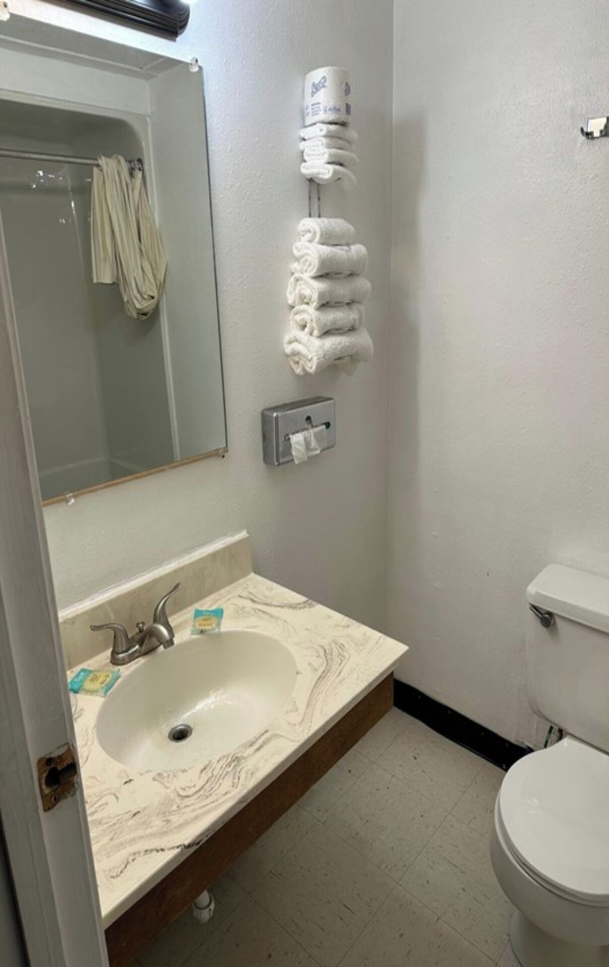 Uma casa de banho com lavatório de mármore, sanita, porta-toalhas com toalhas dobradas, espelho e dispensador de papel.