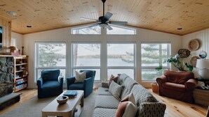 Ein gemütliches Wohnzimmer mit Holzbalkendecke, großen Fenstern, einem Deckenventilator und einem Kamin aus Stein.