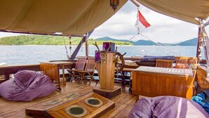 A boat deck with wooden furniture, a steering wheel, and a view of a lake with hills in the background. - Komodo Pirate Boat - Phinisi Liveaboard (Labuan Bajo)