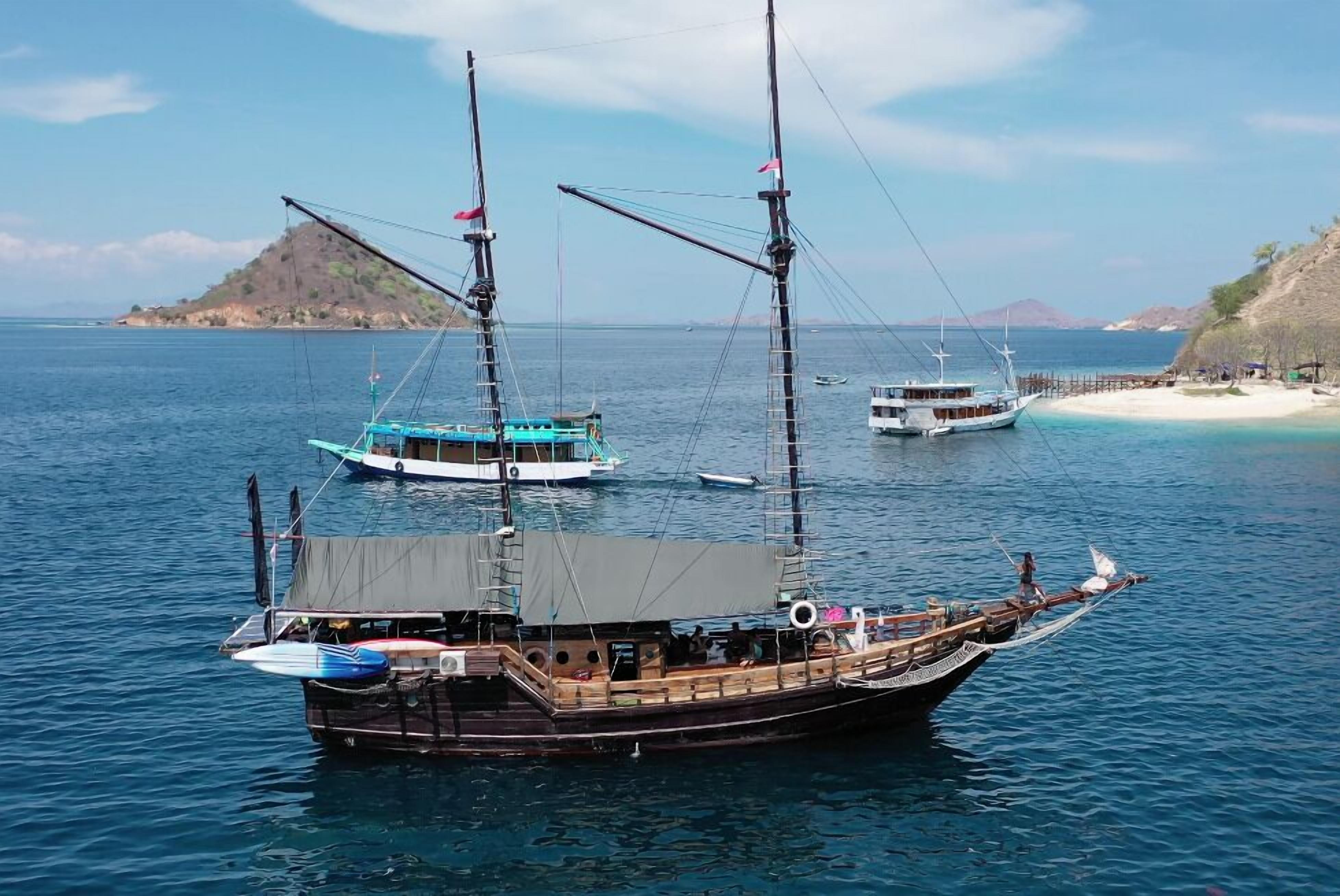A traditional wooden boat with a covered deck, anchored near a sandy beach with a small island in the background.