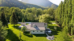 A two-story house with a sloped roof, surrounded by lush greenery and a mountain in the background.