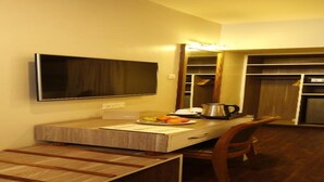 Quarto de hotel compacto com escrivaninha de madeira, televisão de tela plana na parede, pequeno espaço de cozinha com cafeteira e armário embutido.