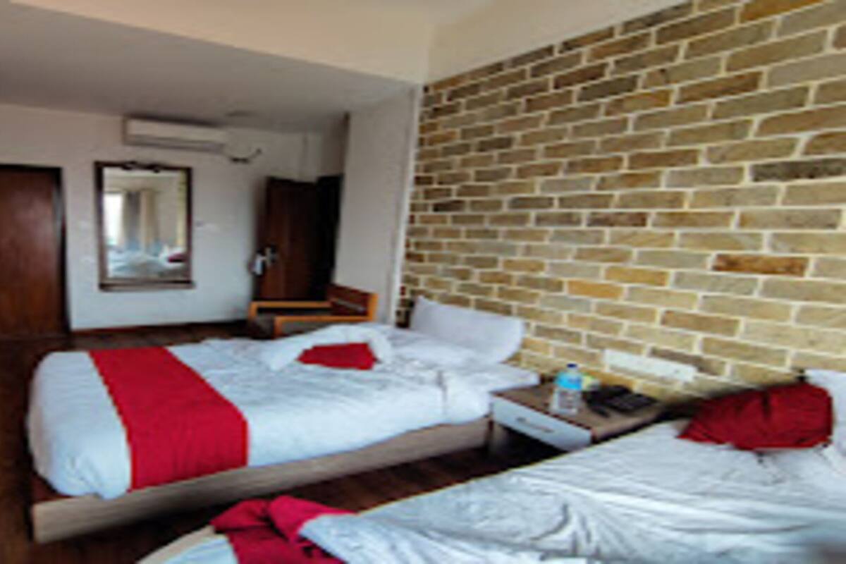 Ein Hotelzimmer mit Backsteinwand, einer Holzbank, zwei Betten mit rot-weißer Bettwäsche, einem kleinen Tisch mit einer Flasche und einem Spiegel an der Wand.
