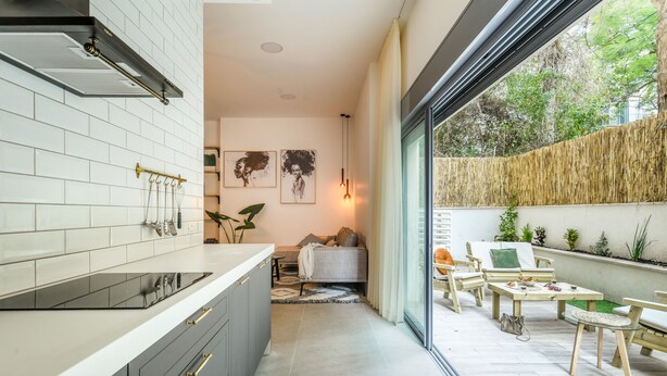 A modern kitchen with white subway tiles, a black induction cooktop, and grey cabinets with gold handles. There is a dining area with outdoor seating and a view of greenery outside. - Spacious 3BR Apt w Garden Ramat Hasharon (Ramat Hasharon)