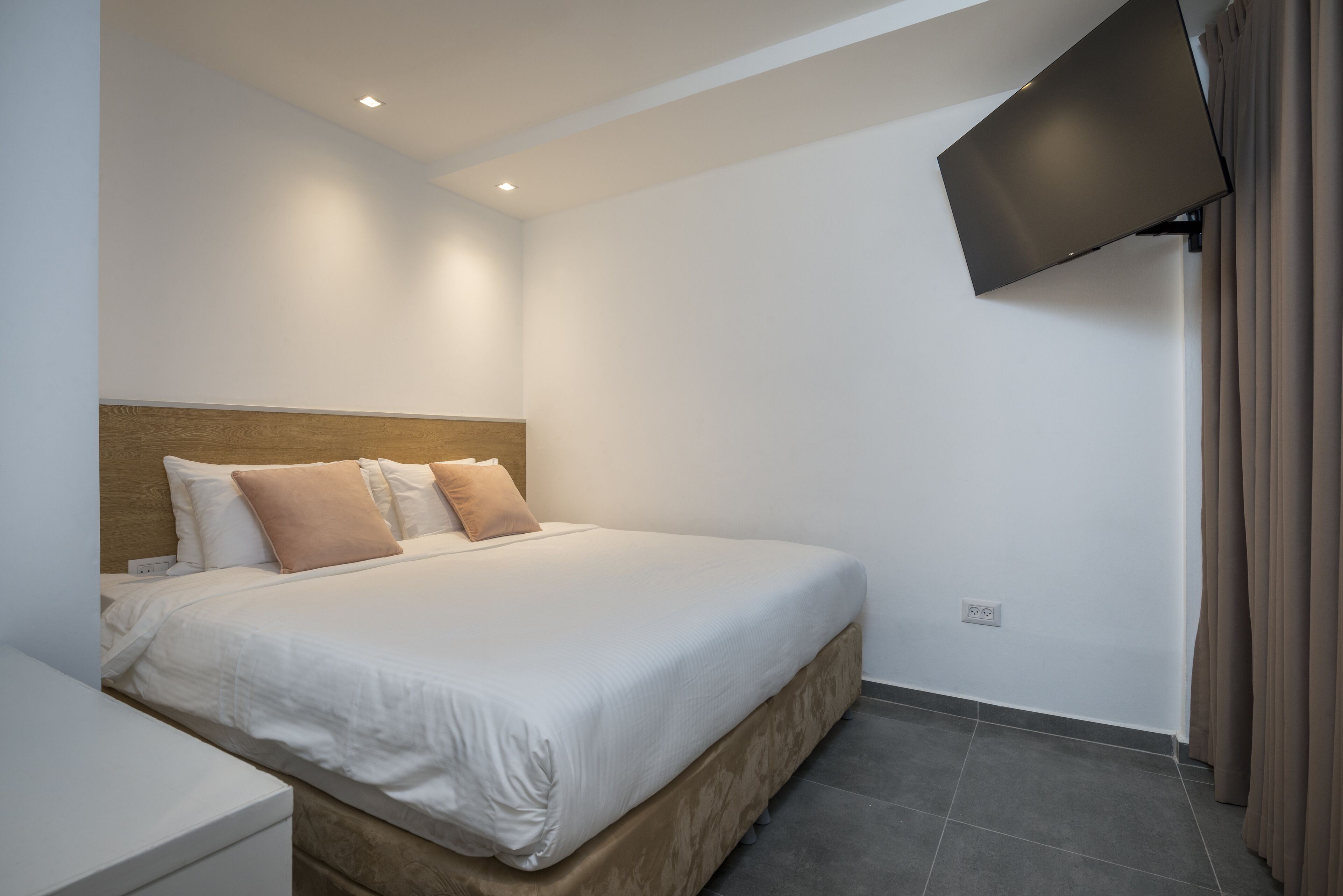 A bedroom with a bed, a bedside table, a flat-screen TV mounted on the wall, and a power outlet on the wall.