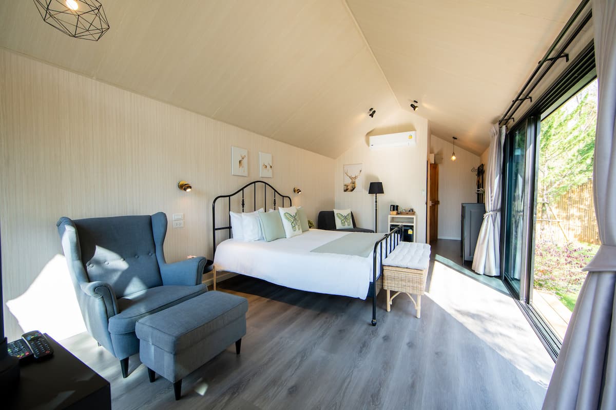 Une chambre moderne avec un grand lit, un coin salon comprenant un fauteuil et un pouf, un bureau avec une télécommande, et une vue sur l’extérieur grâce à de grandes fenêtres.