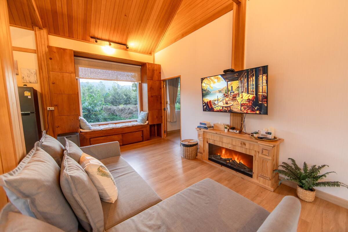 Un salon chaleureux avec un plafond en bois, une cheminée, un grand téléviseur et une fenêtre donnant sur un paysage verdoyant.