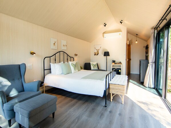 Relax in this serene bedroom with a large bed, armchair, and wooden bench, offering a peaceful retreat with a view of lush greenery.