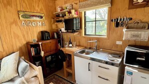 A cozy wooden interior with a sofa, a small dining table, a kitchen area with a sink, stove, and refrigerator, and a window providing natural light. - Prvt cottage + cabin + pool on quarter acre section (wood burner, pet friendly) (Greytown)
