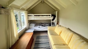 A dorm-style room with bunk beds, a sofa, a wooden bench, and a window with curtains. - Prvt cottage + cabin + pool on quarter acre section (wood burner, pet friendly) (Greytown)