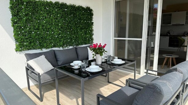 An outdoor dining area with a glass table, grey sofa, and a wall-mounted green plant.