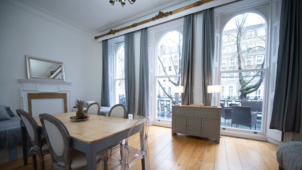 Un'ampia stanza con un tavolo da pranzo in legno, sedie, un divano e un piccolo mobile. Grandi finestre offrono una vista sulla città.