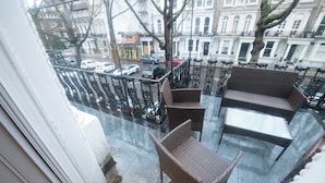 Un balcone con mobili in vimini, un tavolo in vetro e vista su auto parcheggiate e edifici.