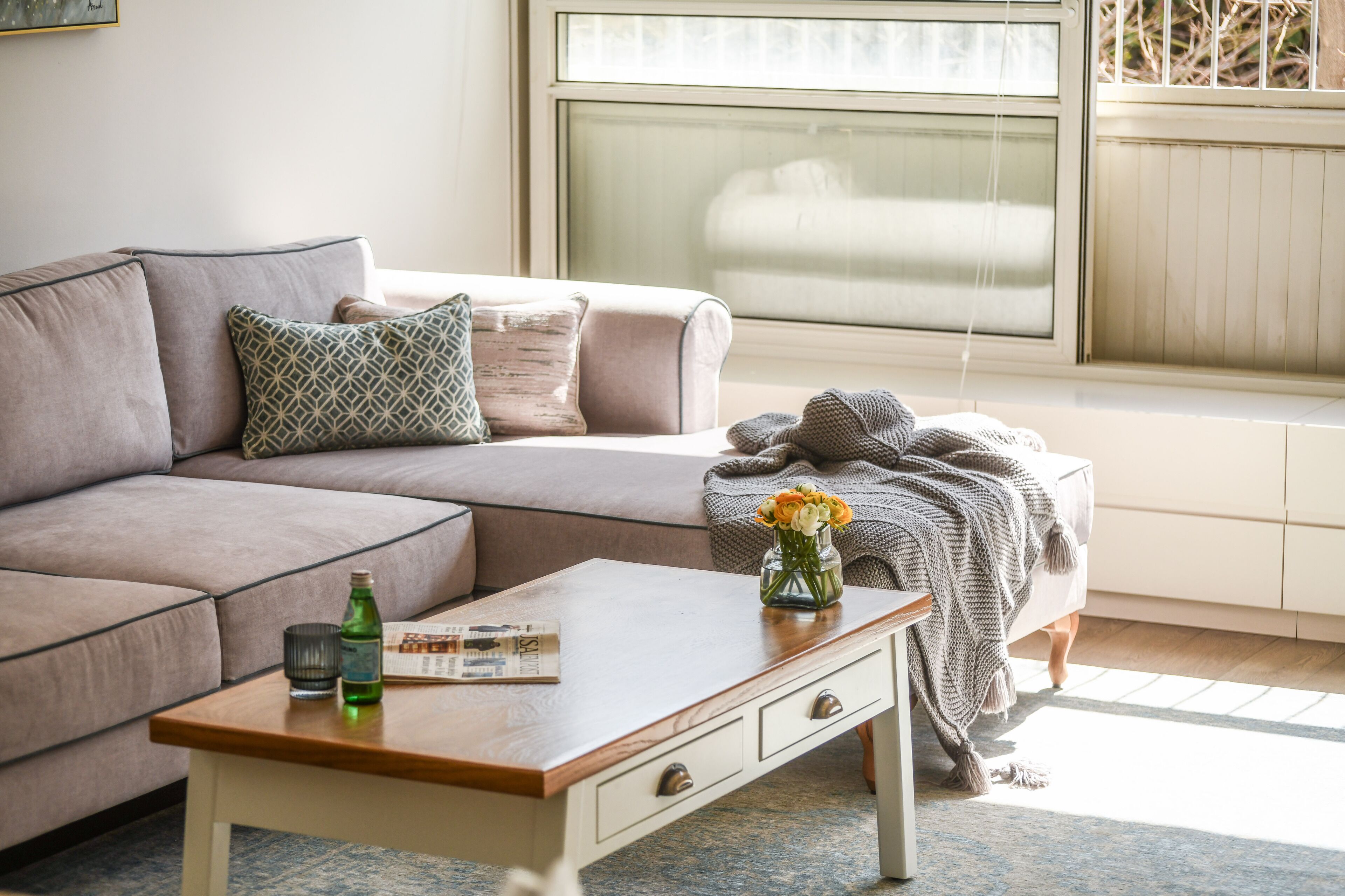 Een woonkamer met een bank, een bijzettafeltje met een bloevas en een raam met jaloezieën.