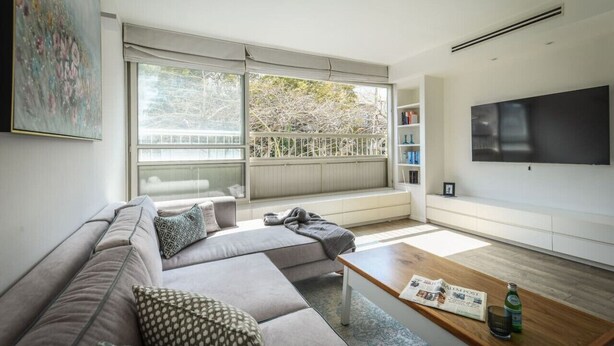 Ein modernes Wohnzimmer mit einem Ecksofa, einem Couchtisch, einem Fernseher und einem großen Fenster mit Blick auf Bäume.