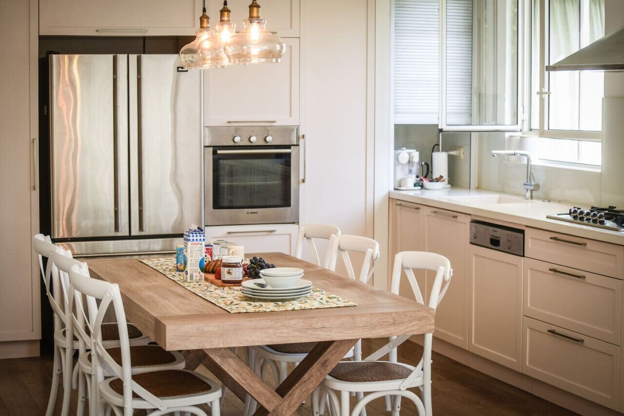 Une cuisine moderne avec une table à manger en bois, des chaises blanches, des appareils électroménagers en acier inoxydable et une fenêtre avec des stores.