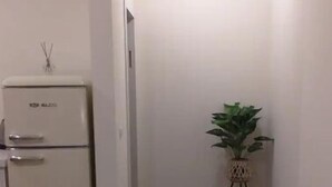 Ein kleiner, schmaler Raum mit einem weißen Kühlschrank, einem Blumentopf auf einem kleinen Tisch und einer Deckenlampe.