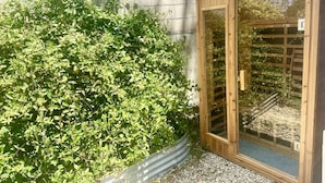 Un cobertizo de madera con puerta de cristal, junto a un matorral verde frondoso, y una pared de metal corrugado al fondo.