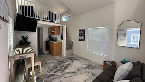 Un salón espacioso con techo alto, una escalera que sube a una buhardilla, un televisor de pantalla plana en la pared y un ventanal grande con persianas.