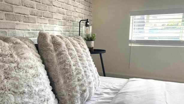 Un dormitorio con pared de ladrillo blanco, cama con ropa de cama blanca y dos almohadas mullidas.