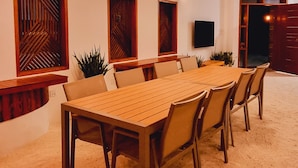 Een eetruimte met een lange houten tafel, meerdere stoelen, plafondventilatoren en wandlampen.