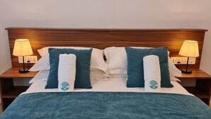Een net opgemaakt bed met een blauwe deken, twee bijpassende kussens en twee bedlampen op houten nachtkastjes.