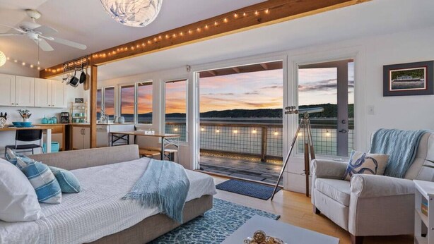 A spacious living area with a large bed, a sofa, and a dining table. There are multiple windows offering a view of the water. - Keys on the Canal - Stunning Beachfront Bungalow! (Belfair)