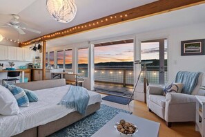 A spacious living area with a large bed, a sofa, and a dining table. There are multiple windows offering a view of the water. - Keys on the Canal - Stunning Beachfront Bungalow! (Belfair)