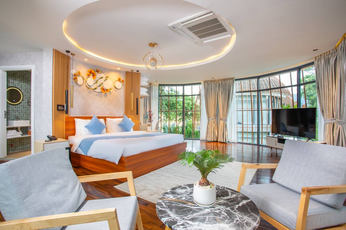A modern hotel room with a large bed, a TV, a plant, and a seating area with a marble table.