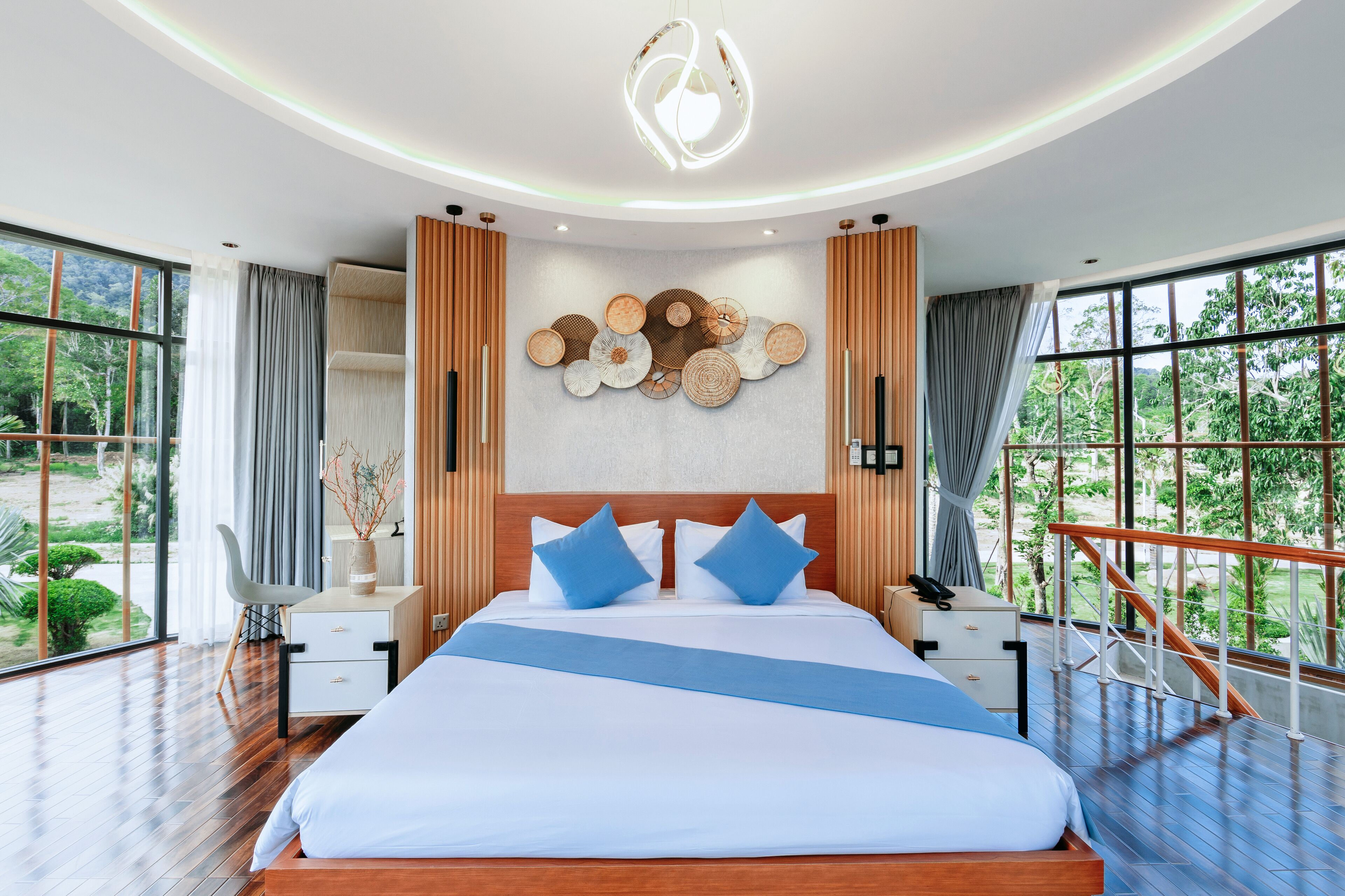 A modern bedroom with a large bed, wooden headboard, and a view of greenery through large windows.