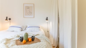 Un dormitorio ordenado con cama, almohadas, una obra de arte enmarcada, un cactus, naranjas y tazas sobre una mesita de noche.