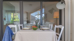 Un comedor con una mesa dispuesta para cuatro personas, sillas blancas, un mantel blanco, un bol de sandía y un jarrón con flores.