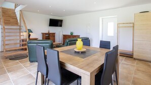 A spacious living area with a wooden dining table, black chairs, a yellow vase, and a TV mounted on the wall.