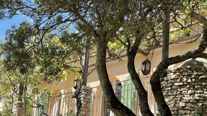 A stone wall with a metal gate, trees, and a building with a stone facade and windows. - Propriété de Charme à Bonifacio (Bonifacio)