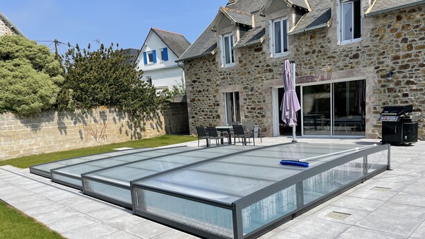 A swimming pool with a glass enclosure, stone house in the background, and outdoor furniture.