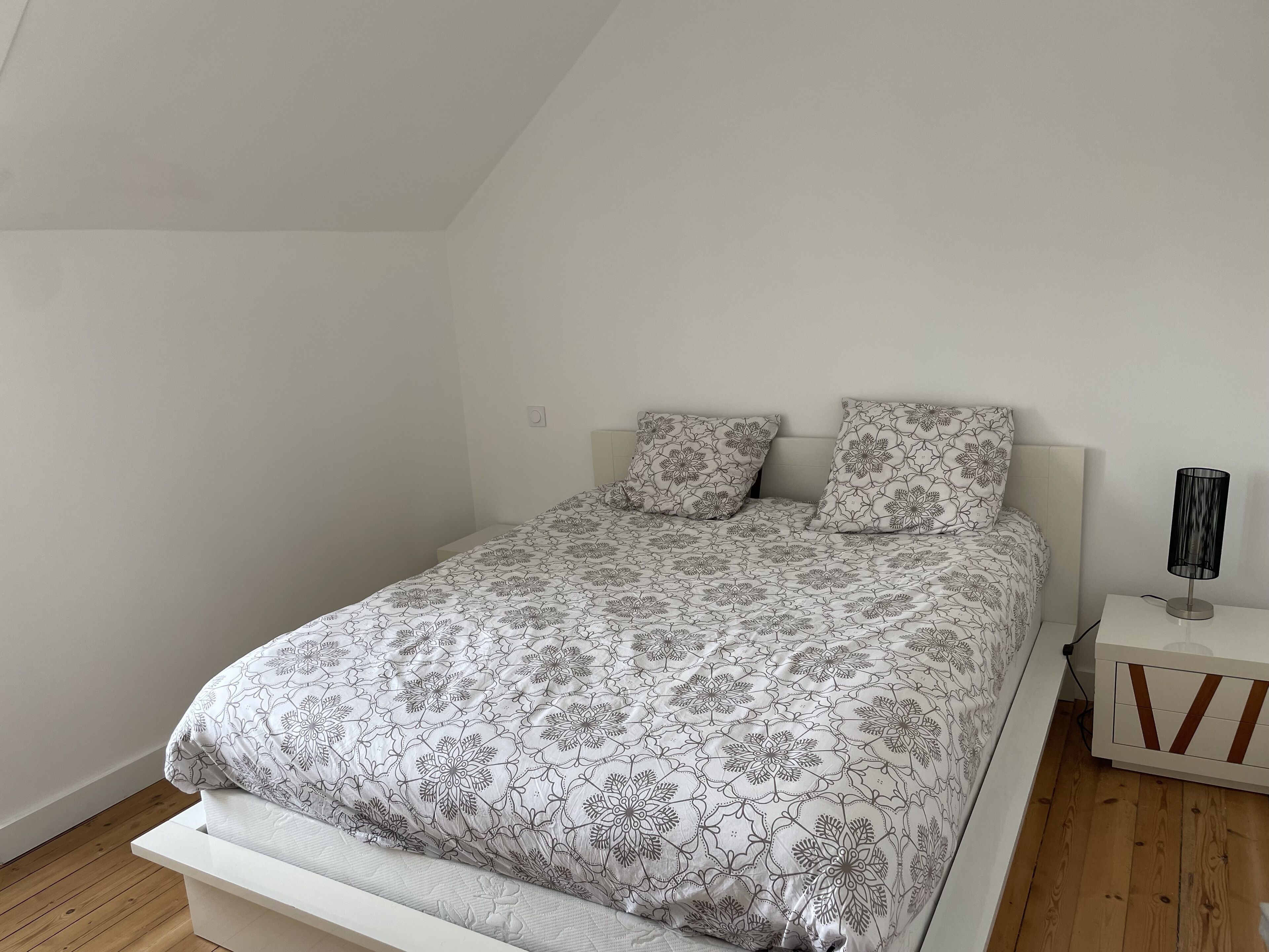 A neatly made bed with a floral-patterned bedspread and pillows, a bedside table with a lamp, and a wooden floor.