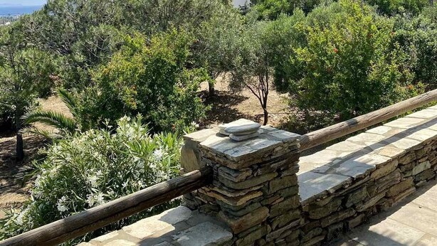 A stone balustrade overlooking a garden with various trees and shrubs, a clear sky, and distant hills. - Cycladic Blue Andros (Γαύριο)