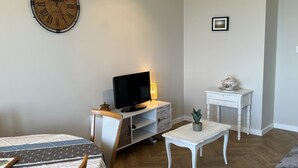 Olohuoneessa on puinen seinäkello, litteänäyttöinen televisio, pieni pöytä, jossa on kasvi, ja valkoinen sivupöytä.