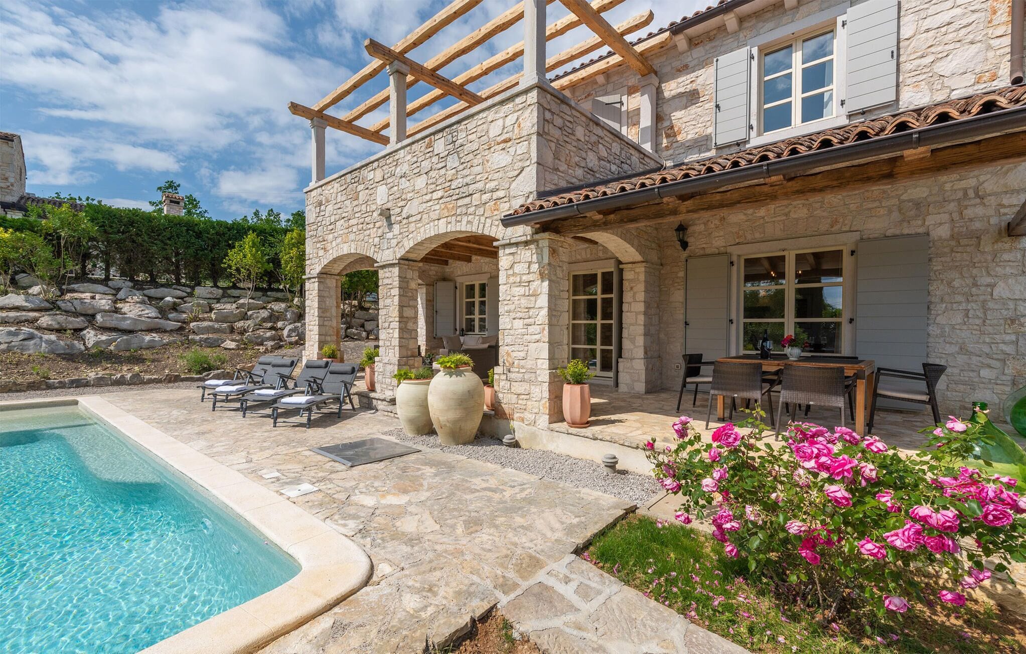 A stone house with a pool, outdoor seating, and a garden with pink flowers.