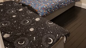 Una habitación pequeña con cama individual, piso oscuro, edredón blanco y negro estampado y otra colcha azul con varios diseños.