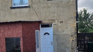 Una casa de dos pisos con fachada roja y color beige, una puerta blanca y dos ventanas. Cuenta con un pequeño patio con dos sillas azules y una mesa.
