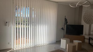 Una sala de estar moderna con un televisor grande, un sofá y un ventanal corredizo con persianas verticales.