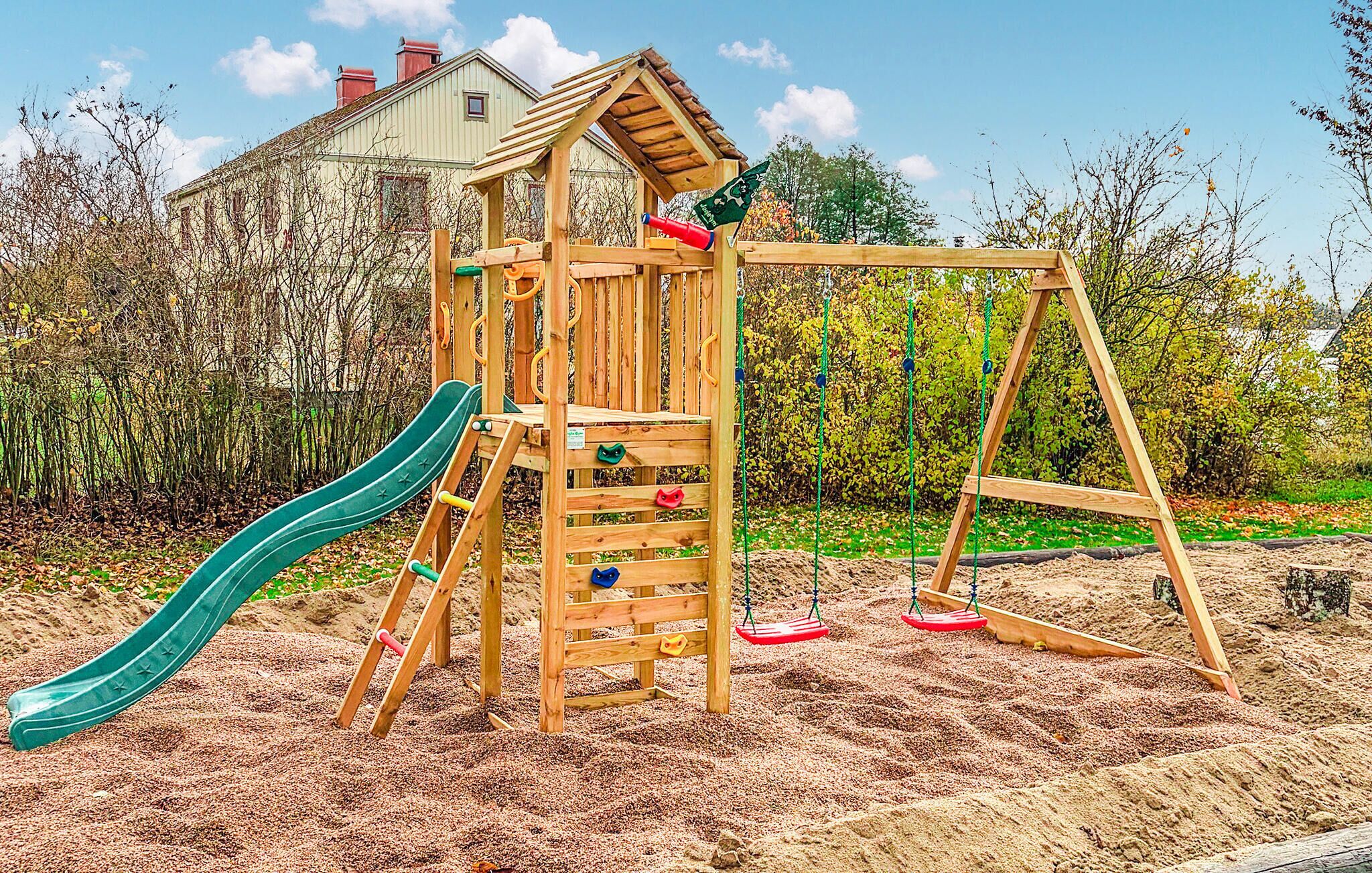 Une structure de jeux en bois comprenant un toboggan, un mur d’escalade et des balançoires, installée dans un espace sablonneux agrémenté d’arbres et avec une maison en arrière-plan.